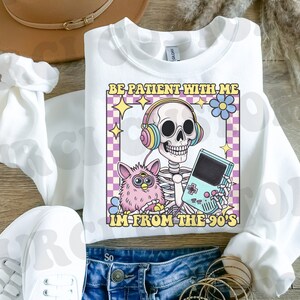 May include: White sweatshirt with a graphic of a skeleton wearing headphones and holding a video game controller. The graphic also includes a pink fuzzy toy and the text "Be patient with me I'm from the 90's".