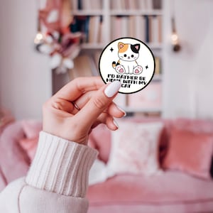 May include: A circular sticker with a white background and a black border featuring a cartoon calico cat with pink paw pads. The text reads I'D RATHER BE HOME WITH MY CAT in black capital letters, accompanied by two small black four-pointed star icons.
