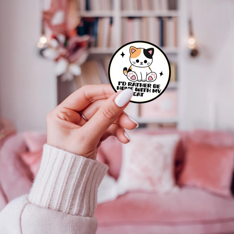May include: A circular sticker with a white background and a black border featuring a cartoon calico cat with pink paw pads. The text reads I'D RATHER BE HOME WITH MY CAT in black capital letters, accompanied by two small black four-pointed star icons.