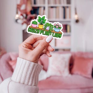May include: A die-cut sticker featuring a wooden shelf holding four potted plants in pink, yellow, purple, and blue pots. Below the shelf, the text CRAZY PLANT LADY is written in bold green letters with a white outline against a white background.