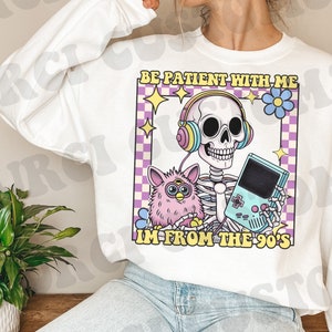 May include: White sweatshirt with a graphic print of a skeleton wearing headphones and holding a video game console. The skeleton is holding a pink fuzzy toy. The text on the graphic says "Be patient with me I'm from the 90's".