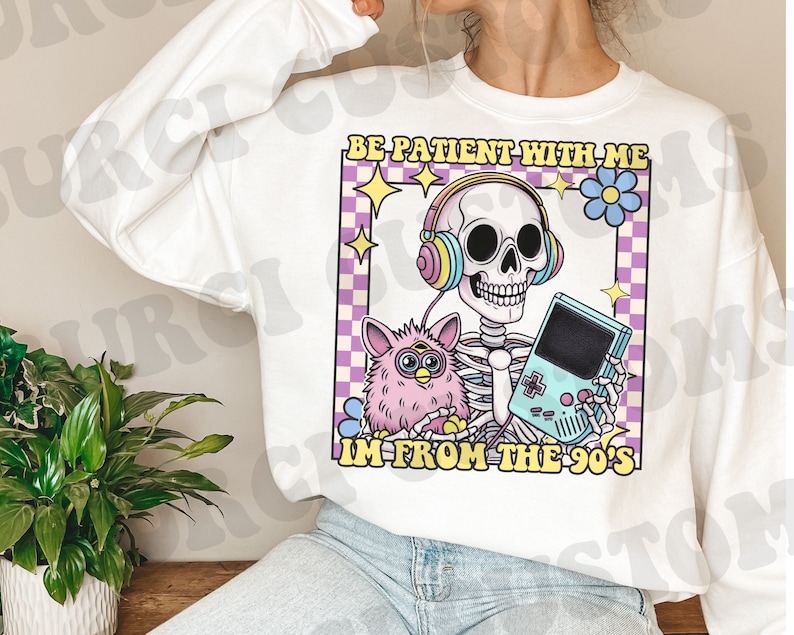 May include: White sweatshirt with a graphic print of a skeleton wearing headphones and holding a video game console. The skeleton is holding a pink fuzzy toy. The text on the graphic says "Be patient with me I'm from the 90's".