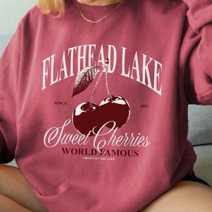 May include: A maroon sweatshirt with a white graphic print of a cherry with the text "Flathead Lake" and "Sweet Cherries World Famous Grown by the Lake" on it.