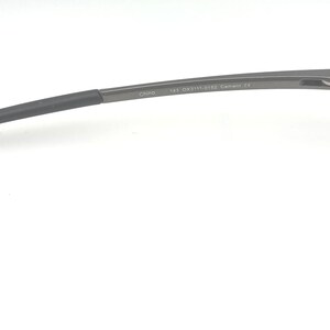 May include: Close-up of a pair of rimless eyeglasses. The temples are dark gray with a curved design, transitioning to a darker, rubberized tip. The frame features a subtle, metallic finish. The lenses are clear.