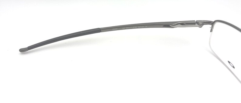May include: Close-up of a pair of rimless eyeglasses. The temples are dark gray with a curved design, transitioning to a darker, rubberized tip. The frame features a subtle, metallic finish. The lenses are clear.