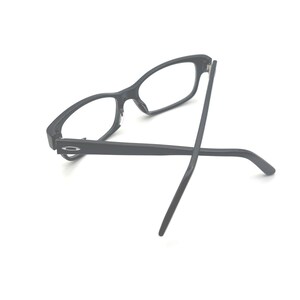 Puede incluir: Gafas rectangulares negras con lentes transparentes. Las gafas tienen un acabado mate y un dise&ntilde;o minimalista. Las patillas son finas y se extienden hacia afuera. Las gafas est&aacute;n sobre un fondo blanco.