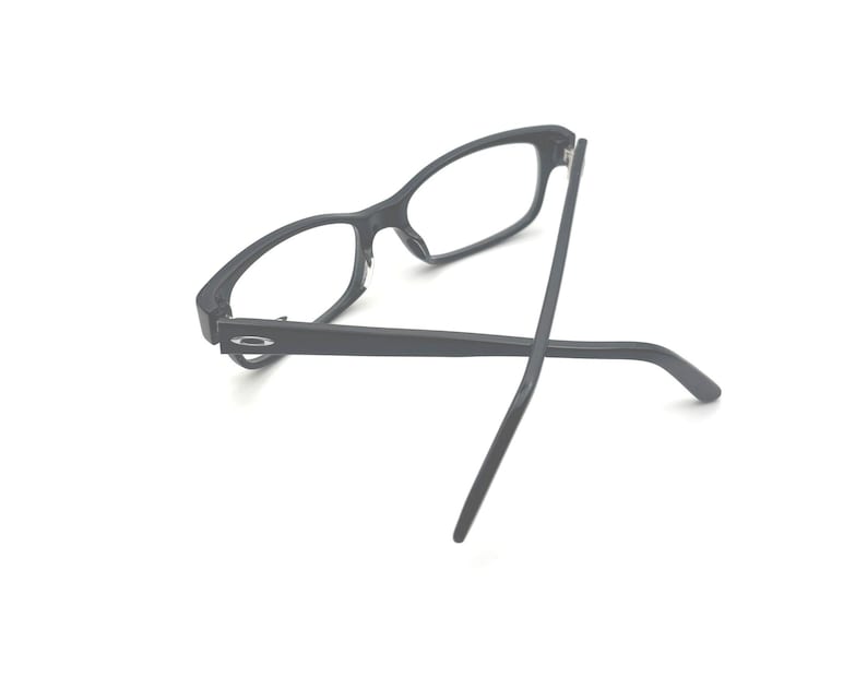 Puede incluir: Gafas rectangulares negras con lentes transparentes. Las gafas tienen un acabado mate y un dise&ntilde;o minimalista. Las patillas son finas y se extienden hacia afuera. Las gafas est&aacute;n sobre un fondo blanco.