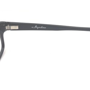 Puede incluir: Gafas negras de forma rectangular, con un dise&ntilde;o minimalista. Las finas patillas llevan el nombre de la marca "Anywhere" en plata. El fondo blanco resalta su elegancia sencilla y discreta.