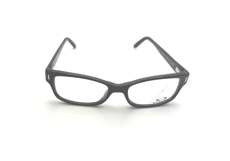 Puede incluir: Gafas grises oscuras con montura rectangular y lentes transparentes. Las gafas tienen una ligera curva en la parte superior de la montura y un peque&ntilde;o logotipo en la lente. Las patillas son finas y se extienden hacia las orejas.