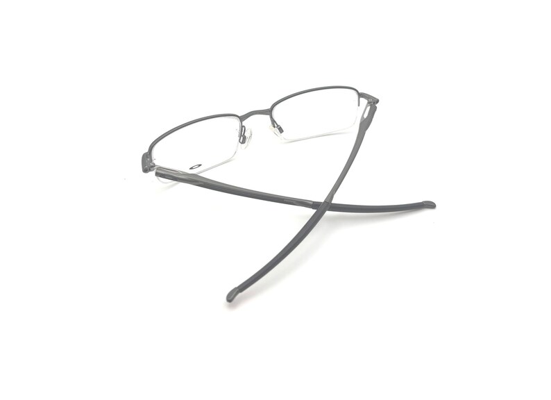 May include: A pair of eyeglasses with a dark gray metal frame and clear lenses. The glasses are set against a white background, with the temples extended. The design is minimalist and modern.