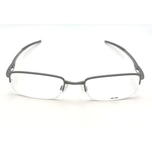 May include: Gray half-rim eyeglasses with clear lenses. The glasses have a rectangular shape at the bottom and a thin, curved frame. The temples are dark gray and extend to the ear pieces. The Oakley logo is visible on the lens.