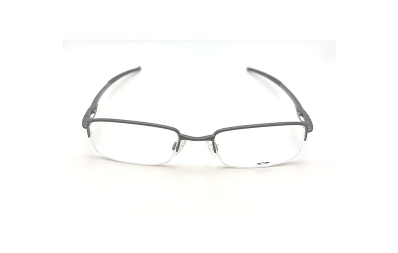 May include: Gray half-rim eyeglasses with clear lenses. The glasses have a rectangular shape at the bottom and a thin, curved frame. The temples are dark gray and extend to the ear pieces. The Oakley logo is visible on the lens.
