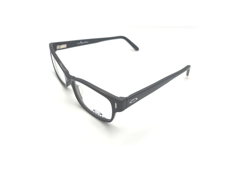 Puede incluir: Gafas rectangulares negras con lentes transparentes. Las gafas tienen un acabado mate y el nombre de la marca es visible en la montura. Las patillas de las gafas son finas y se extienden hacia atr&aacute;s.