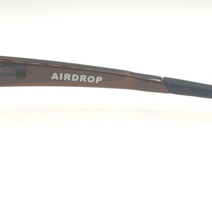 May include: Close-up of a pair of eyeglasses with a brown frame and black arm. The text "AIRDROP" is printed on the arm. The glasses are set against a white background.