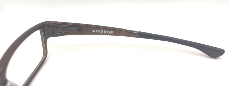 May include: Close-up of a pair of eyeglasses with a brown frame and black arm. The text "AIRDROP" is printed on the arm. The glasses are set against a white background.