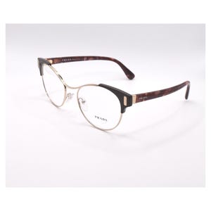 May include: Gold-rimmed eyeglasses with a tortoiseshell frame. The frame has a subtle cat-eye shape and the brand name "PRADA" is printed on the right temple.