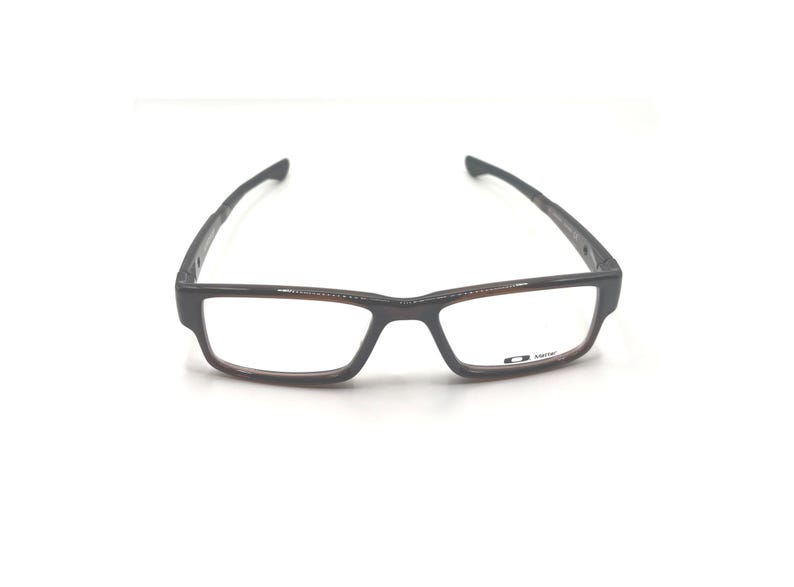 May include: Rectangular eyeglasses with a dark gray frame and brown accents. The lenses are clear, and the temples are thin and angled. The brand logo is visible on the lower right lens.