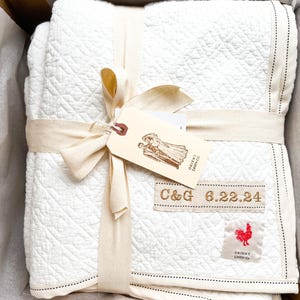 May include: A white quilted blanket with a cream ribbon tied around it. The blanket has a black stitched border and a small red tag with a rooster logo and the text "Orient Linen Co.". The tag is attached to a larger cream tag with the text "C&G 6.22.24" and a small illustration of a couple.