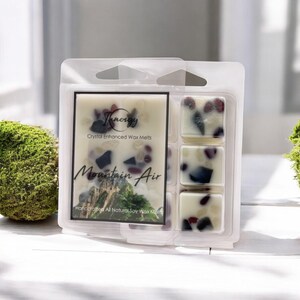 May include: A package of six soy wax melts with a mountain air scent. The wax melts are infused with crystals and are handcrafted. The package is white with black text that reads "Innergy Crystal Enhanced Wax Melts Mountain Air Handcrafted All Natural Soy Wax Melts".