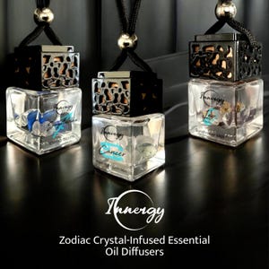 May include: Three glass car air fresheners with black metal tops and zodiac signs. The signs are Sagittarius, Cancer, and Leo. Each bottle contains crystals and essential oils. The text on the bottles reads "Innergy" and the scent names. The text at the bottom of the image reads "Zodiac Crystal-Infused Essential Oil Diffusers".