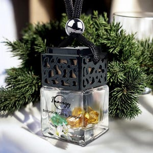 Op de afbeelding: Een heldere, vierkante glazen luchtverfrisser met de tekst "Innergy Botanical Gin & Juniper". De luchtverfrisser heeft een zwarte, patroonvormige bovenkant en bevat kleurrijke kristallen en witte bloemen. Een zwart koord met een zilveren bal hangt van bovenaf.