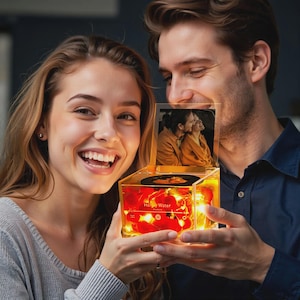 Puede incluir: Una caja transparente e iluminada con una foto y un pequeño disco en el interior. La caja está llena de elementos decorativos rojos y luces de hadas. La caja está sostenida por dos personas, con una foto de una pareja en la parte superior.