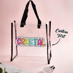 Peut inclure: Un sac fourre-tout transparent avec des bordures et des poignées noires. Le sac affiche le nom "CRYSTAL" en lettres colorées. Le fond est rose clair.