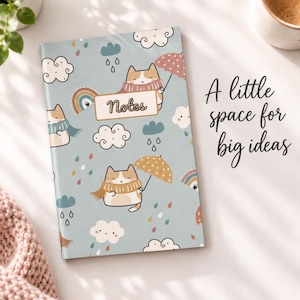 May include: A light blue notebook featuring a repeating pattern of cats with scarves and umbrellas, clouds, rainbows, and raindrops. A central label reads Notes. The text A little space for big ideas is printed to the right of the notebook.