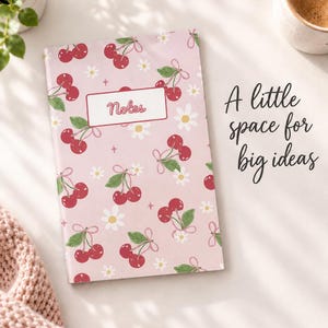May include: A rectangular notebook with a light pink cover featuring a repeating pattern of red cherries, green leaves, and white daisies. A white rectangular label in the center contains the word Notes. The text A little space for big ideas is visible.