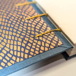 May include: A handmade journal with a blue denim cover and a patterned cover with gold and blue details. The pages are white and the binding is secured with yellow thread.