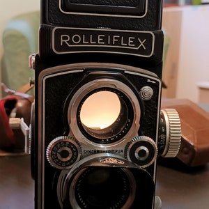 以下が含まれることがあります： 黒と銀の Rolleiflex 双眼鏡式カメラと薄い茶色の革ケース。カメラは大きなファインダーと銀色のリングが付いたレンズを備えています。カメラは暗い表面の上にあります。