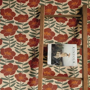 May include: Floral wallpaper featuring a repeating pattern of large, stylized red flowers with yellow centers and green leaves on a cream background. A wooden ladder leans against the wall, with a magazine titled "KINFOLK" resting on a rung.