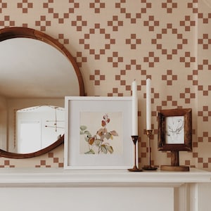 May include: A white mantelpiece with a brown wooden mirror, two white picture frames, and two brass candle holders with white candles. The wall behind the mantelpiece is covered in a brown and beige geometric wallpaper.