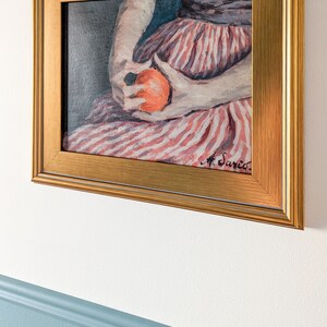 Painting of a woman holding fruit in a gold frame on a white wall