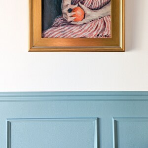 Painting of a woman holding fruit in a gold frame on a white wall