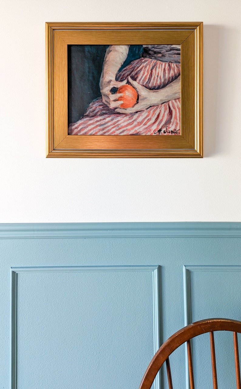 Painting of a woman holding fruit in a gold frame on a white wall