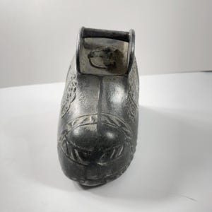 Op de afbeelding: Een donkergrijs metalen schoenvormig object met een handvat. De schoen heeft sierlijke bloemen en geometrische patronen. De handgreep is een gebogen staaf die aan de bovenkant is bevestigd. De schoen heeft een klein paardenkopontwerp aan de bovenkant.