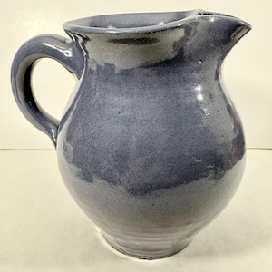 May include: A handmade, blue ceramic pitcher with a rounded body, a curved handle, and a pouring spout. The pitcher has a glossy finish and a slightly irregular shape, suggesting it is handcrafted.