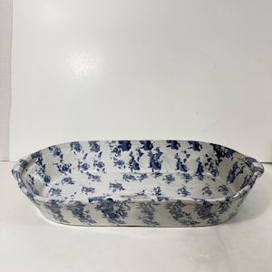 May include: An oval-shaped ceramic baking dish with a white base and a blue floral pattern. The dish has a slightly raised rim and handles on each end. The design is reminiscent of vintage kitchenware.