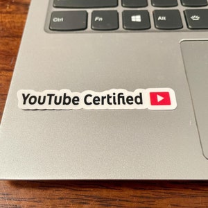 Może przedstawiać: Srebrny laptop z czarną klawiaturą i białą naklejką z napisem "YouTube Certified" z czerwoną ikoną przycisku odtwarzania.