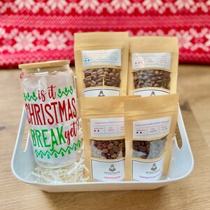 May include: A white gift basket filled with white shredded paper and four bags of coffee beans. The bags are labeled with the text "The Bad Teacher Coffee Roasters" and the names of the coffee blends: "Maestra Blend" and "Surprise Facets". The basket also contains a clear glass jar with a bamboo lid and a festive Christmas-themed sticker that reads "Is it Christmas Break yet?"
