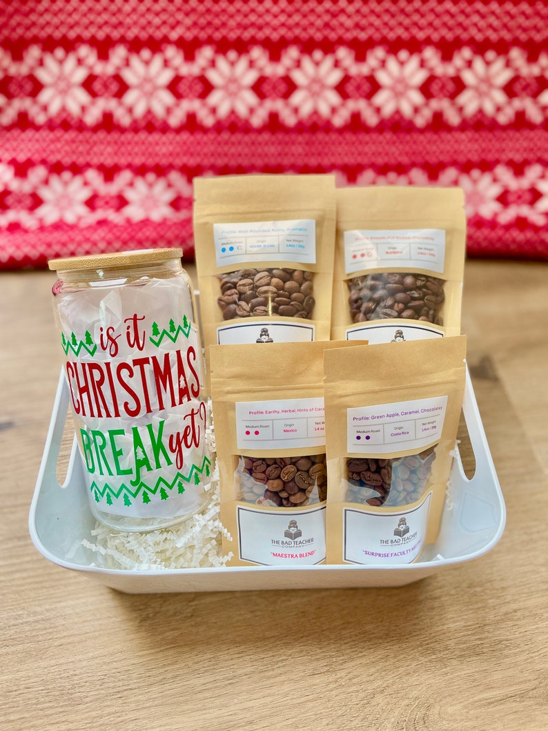 May include: A white gift basket filled with white shredded paper and four bags of coffee beans. The bags are labeled with the text "The Bad Teacher Coffee Roasters" and the names of the coffee blends: "Maestra Blend" and "Surprise Facets". The basket also contains a clear glass jar with a bamboo lid and a festive Christmas-themed sticker that reads "Is it Christmas Break yet?"