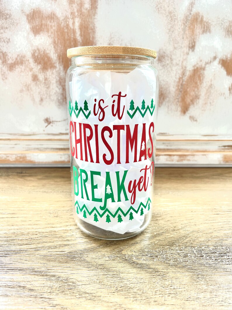 May include: A clear glass jar with a bamboo lid and a red and green Christmas themed design that says "is it Christmas break yet?"