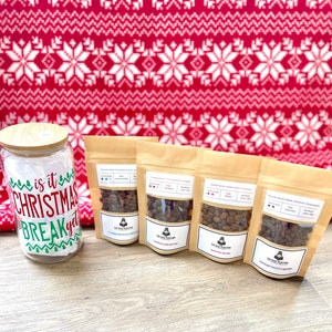 May include: Four bags of coffee beans with different flavors:  "Clandestine Walk Through", "Season Plans Are Me", "Salestra Blend", and "Swish Falls Mystic". The bags are on a wooden surface with a glass jar with the text "Is it Christmas Break yet?" in the background.