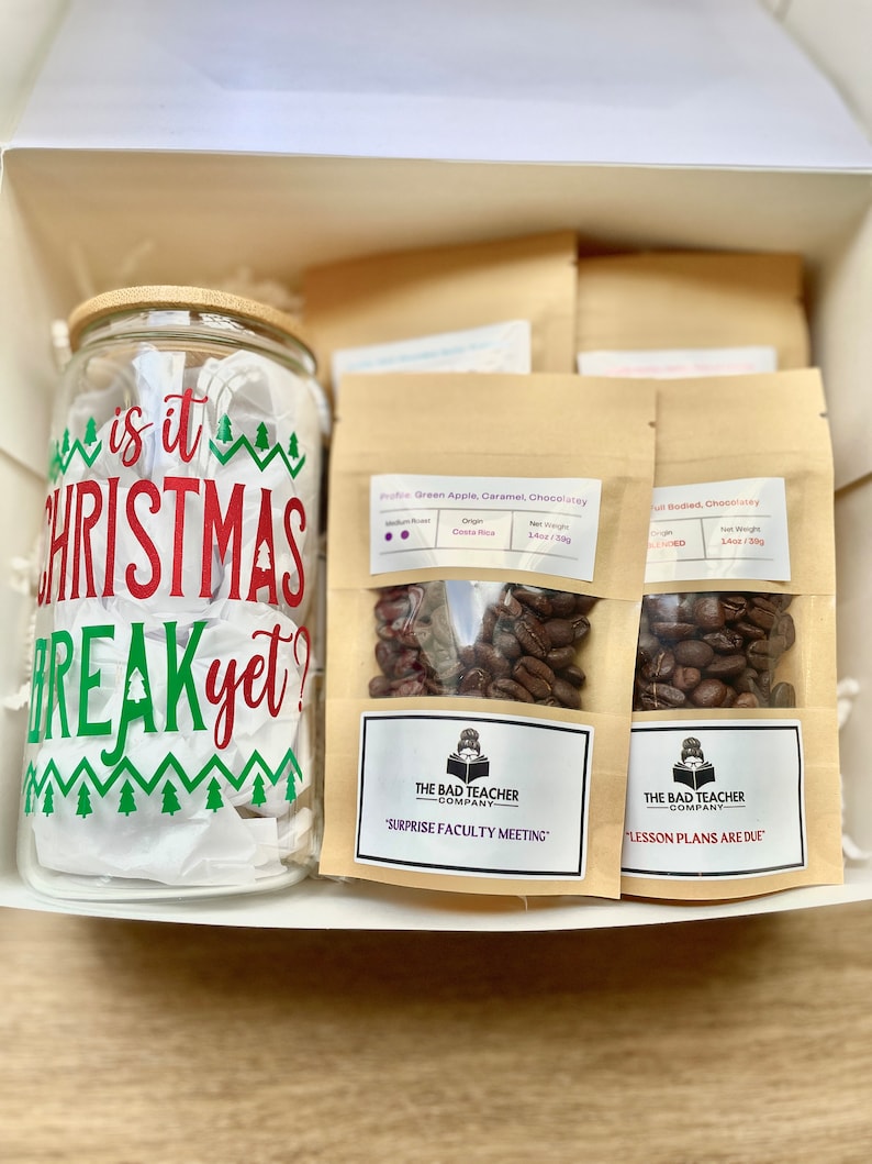 May include: A gift box filled with three bags of coffee beans and a glass jar with a Christmas-themed design that says "Is it Christmas break yet?" The coffee bags are labeled with the text "The Bad Teacher Company" and the text "Surprise Faculty Meeting" and "Lesson Plans Are Due".
