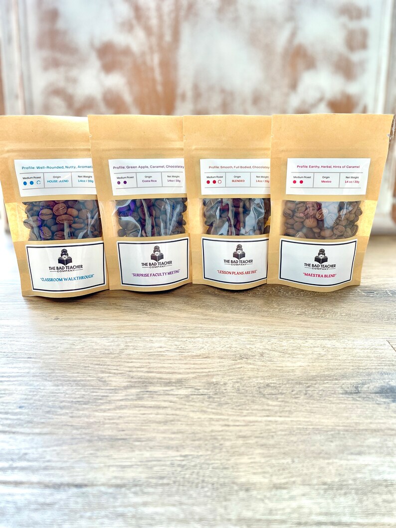 May include: Four brown paper bags with clear windows containing roasted coffee beans. Each bag has a label with the text "The Bad Teacher Company" and a different blend name: "Classroom Walkthrough", "Surprise Faculty Meeting", "Lesson Plans Are Due", and "Maestra Blend".