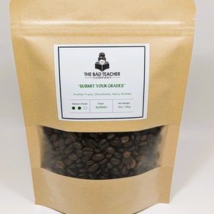 Pode incluir: Um saco de papel castanho com uma janela transparente que mostra grãos de café torrados. O saco tem um rótulo branco com o texto "THE BAD TEACHER COMPANY" e "SUBMIT YOUR GRADES". O rótulo também inclui informações sobre o torrado, a origem e o peso líquido: Torrado médio, Mistura, 141 g.