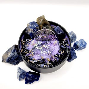 Puede incluir: Una vela morada con un amuleto plateado en el centro, rodeada de piedras negras y azules. La vela est&aacute; en un recipiente de hojalata negro.