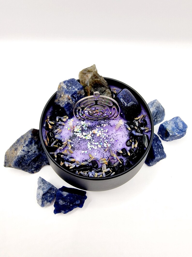 Puede incluir: Una vela morada con un amuleto plateado en el centro, rodeada de piedras negras y azules. La vela est&aacute; en un recipiente de hojalata negro.