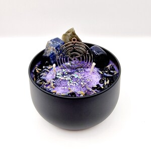 Puede incluir: Un tarro de vela negro con una parte superior de cera morada. El tarro contiene cristales, purpurina y un amuleto de metal. El amuleto es un c&iacute;rculo con un dise&ntilde;o geom&eacute;trico.